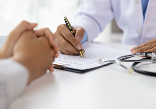 A professional in a white coat takes notes opposite a person with clasped hands, with a stethoscope on the desk.
