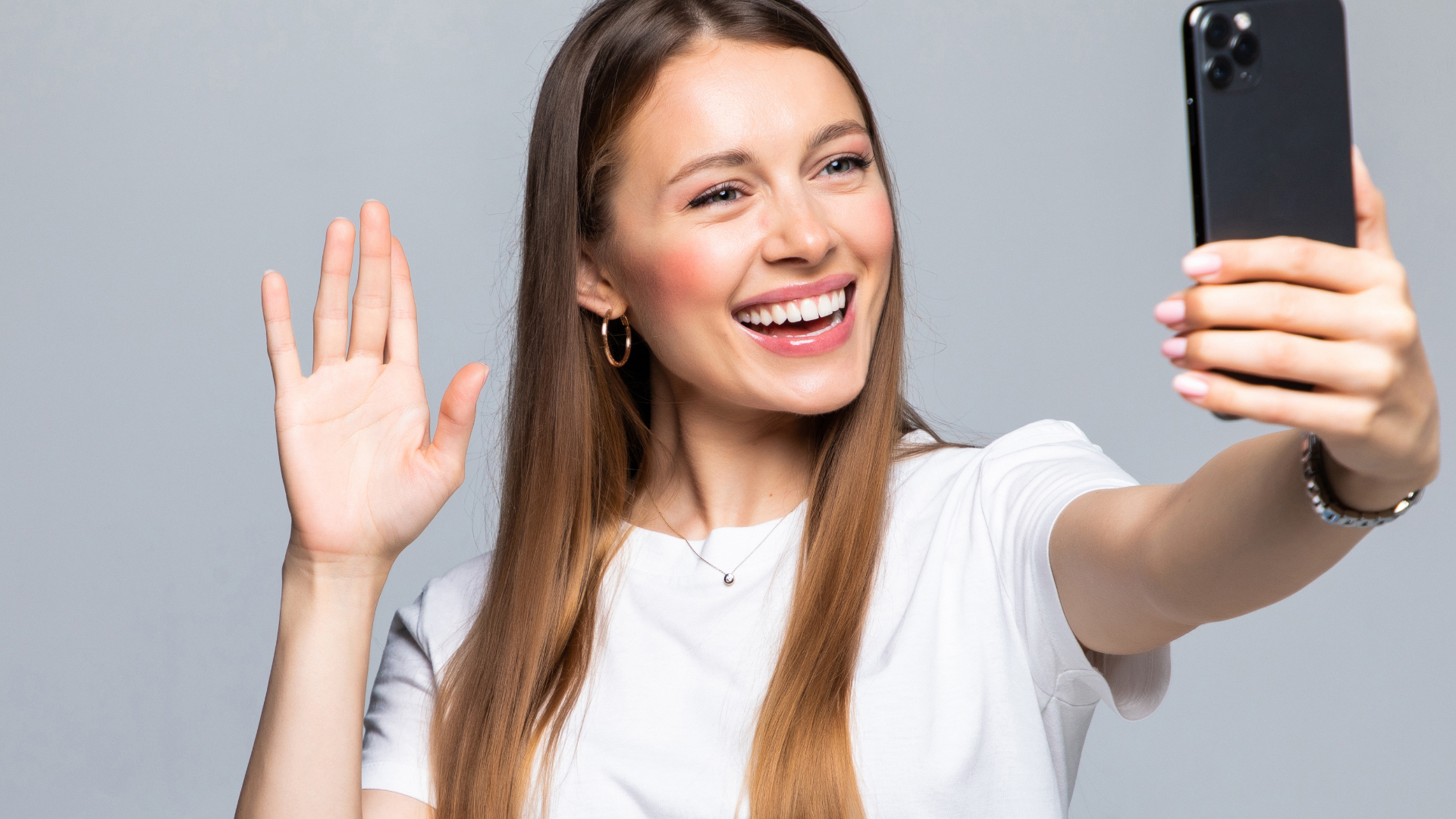 A young woman in a white t-shirt waving and smiling broadly while taking a selfie with her phone