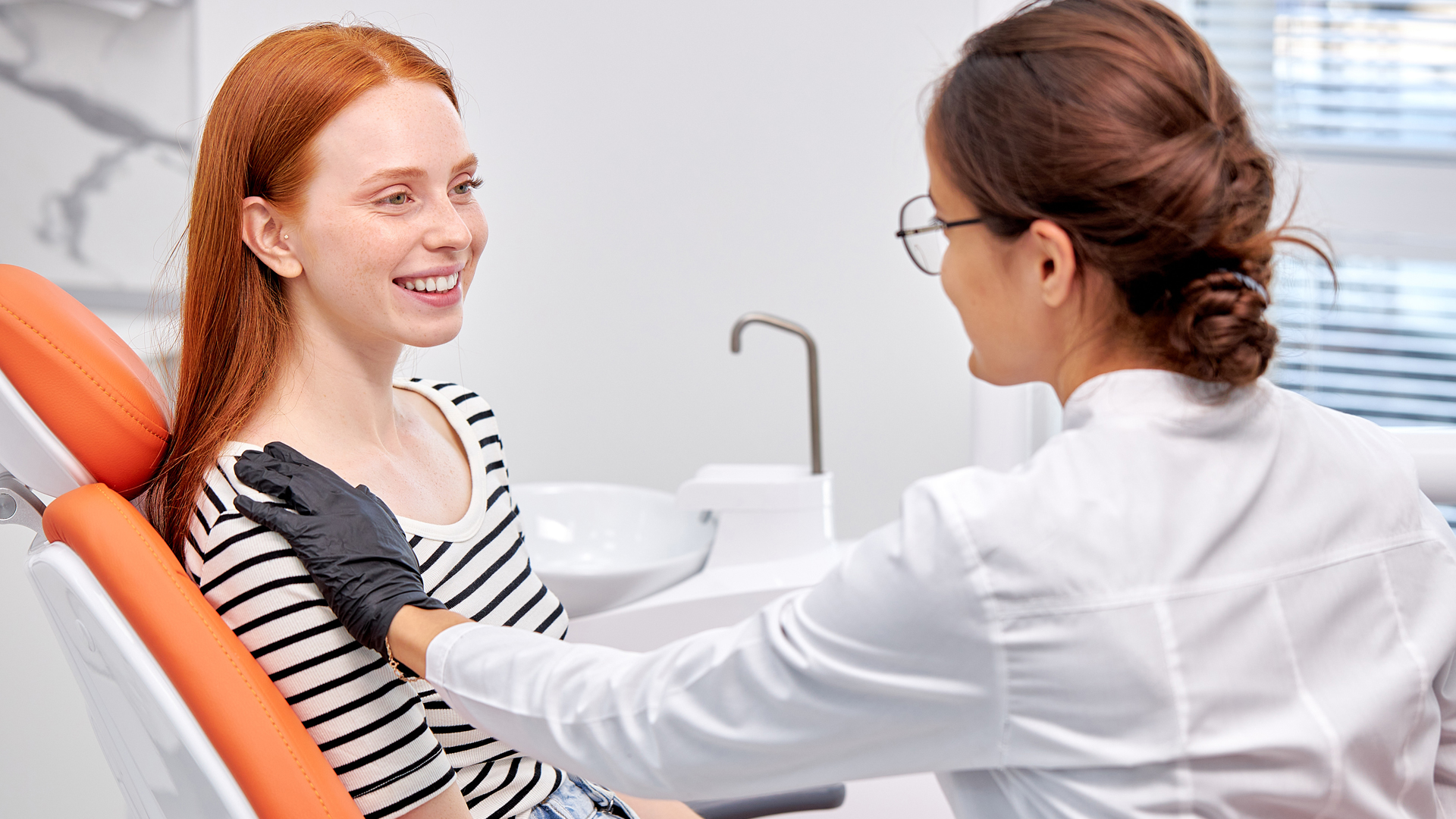 Young red-haired patient sitting in the chair, smiling, talking with a dentist in a white coat