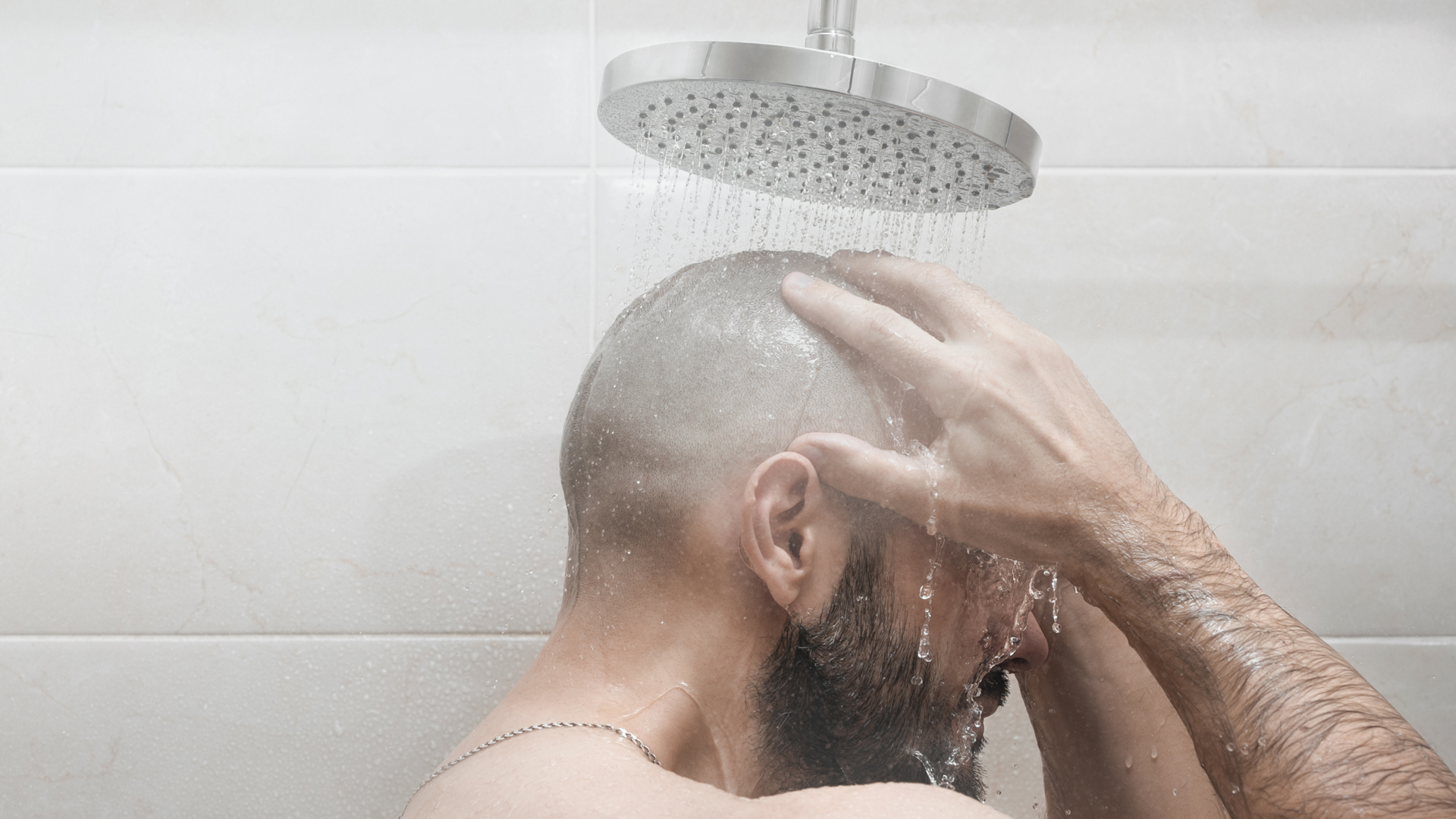 Homme au crâne rasé sous un pommeau de douche, une main posée sur la tête tandis que l’eau coule.