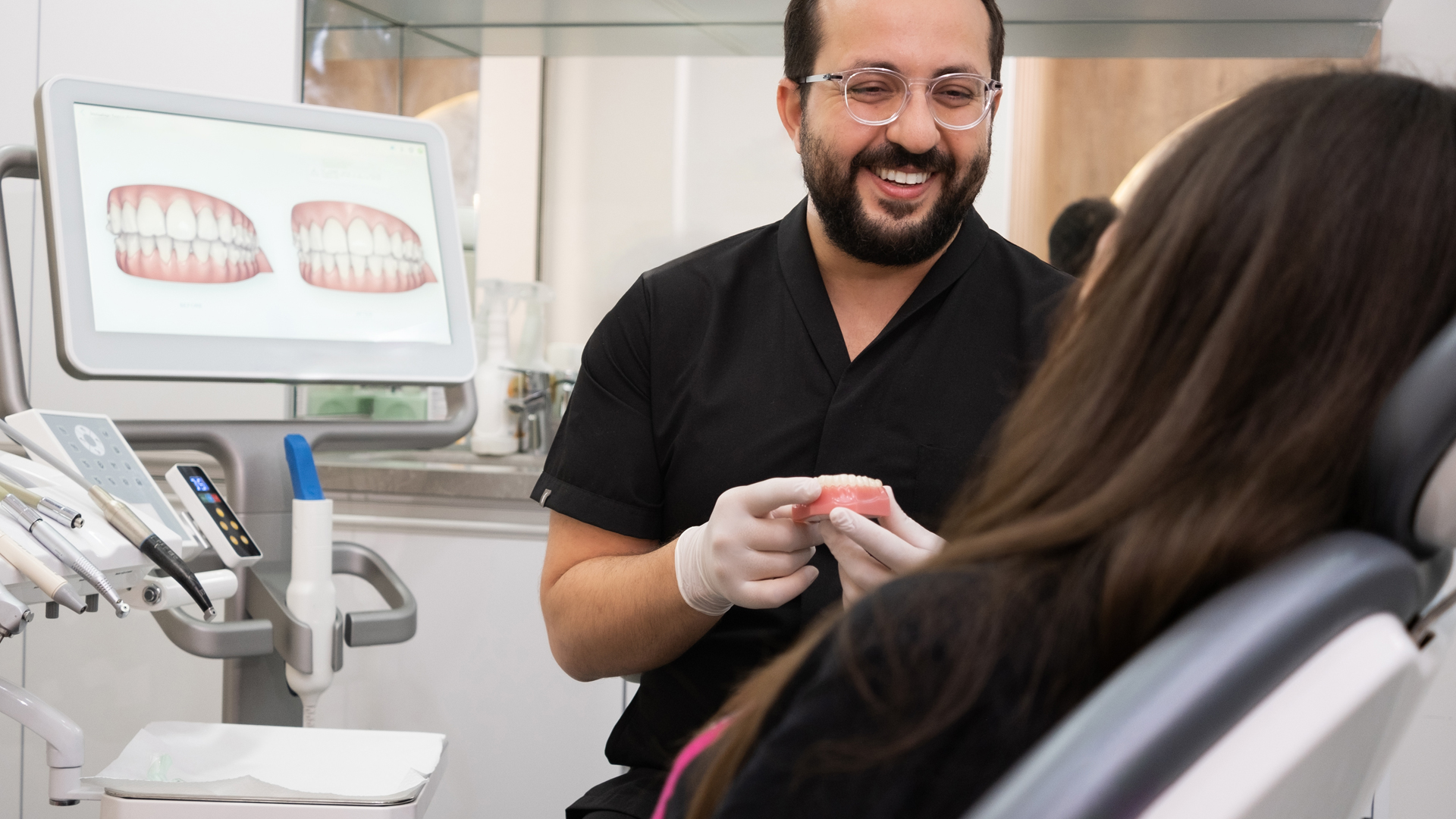 Smiling dentist holding a dental model in front of a seated patient, with images of teeth displayed on a screen beside them