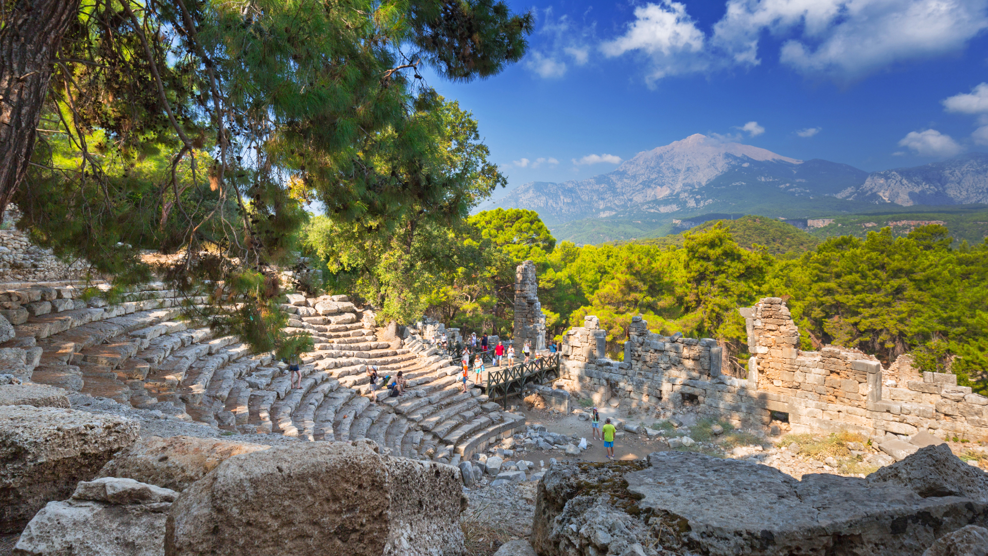 Stone tiers of an ancient theatre set in a pine forest, with the peaks of the Taurus Mountains behind