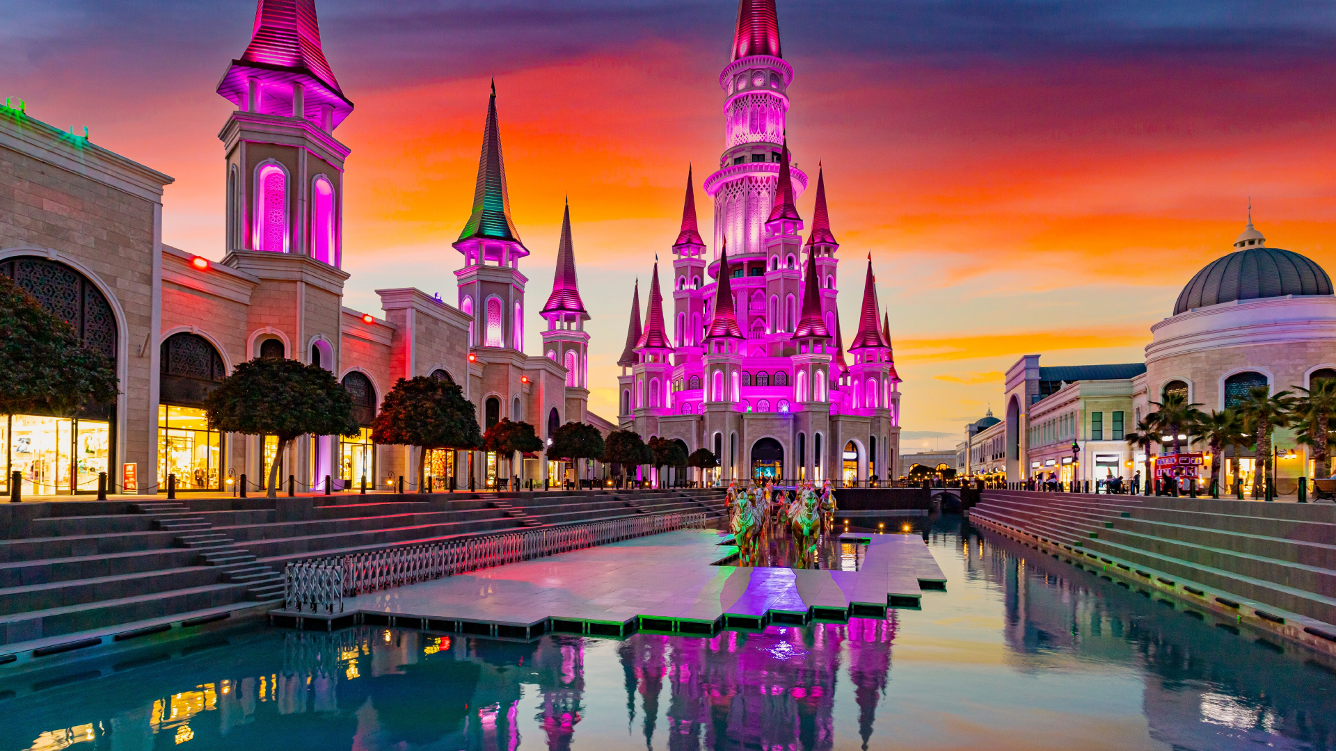 Floodlit theme park near Antalya at dusk, a pink-lit castle reflected in a canal lined with shops