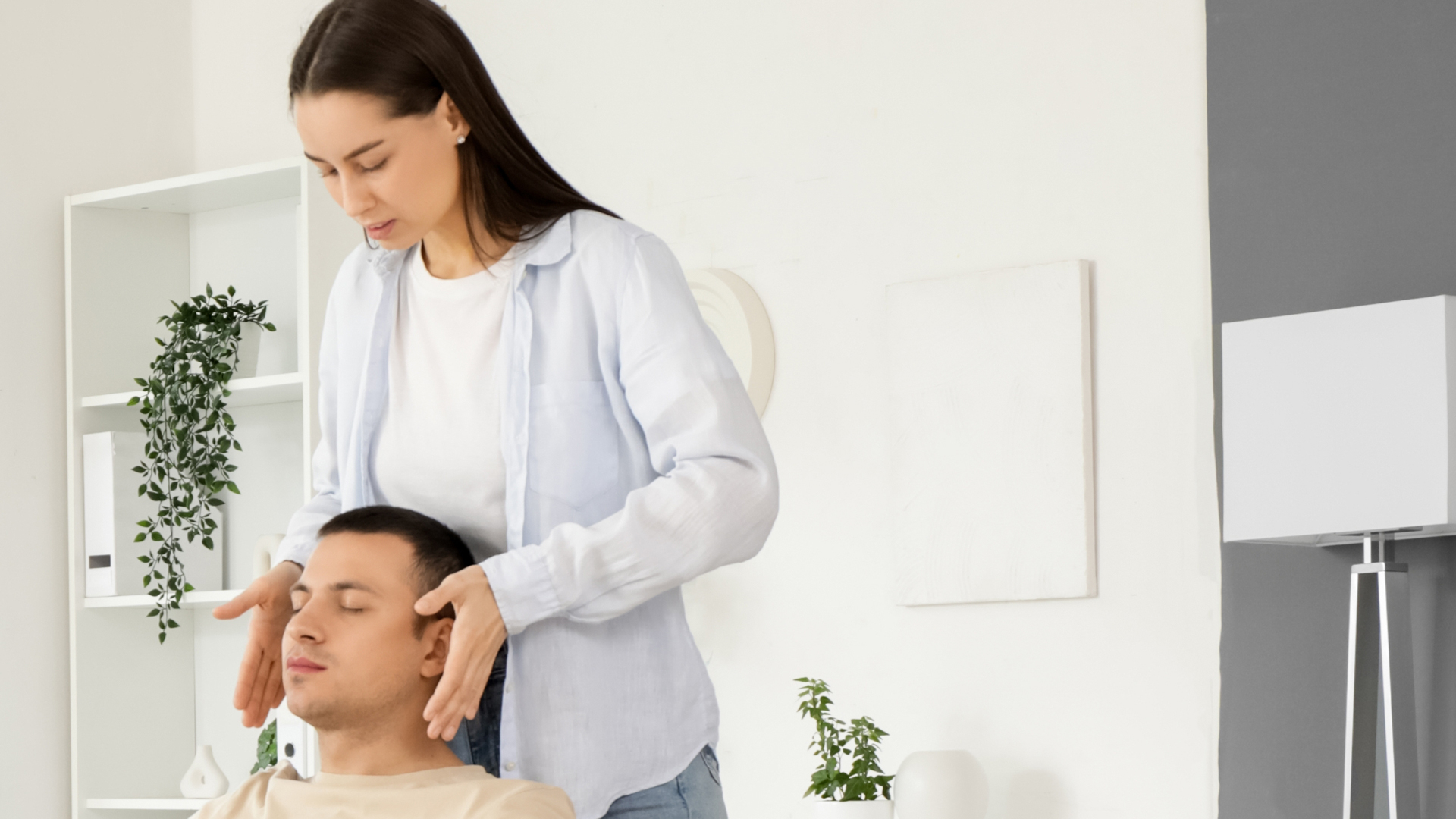 Practitioner standing behind a man lying with his eyes closed, hands either side of his head, in a bright room