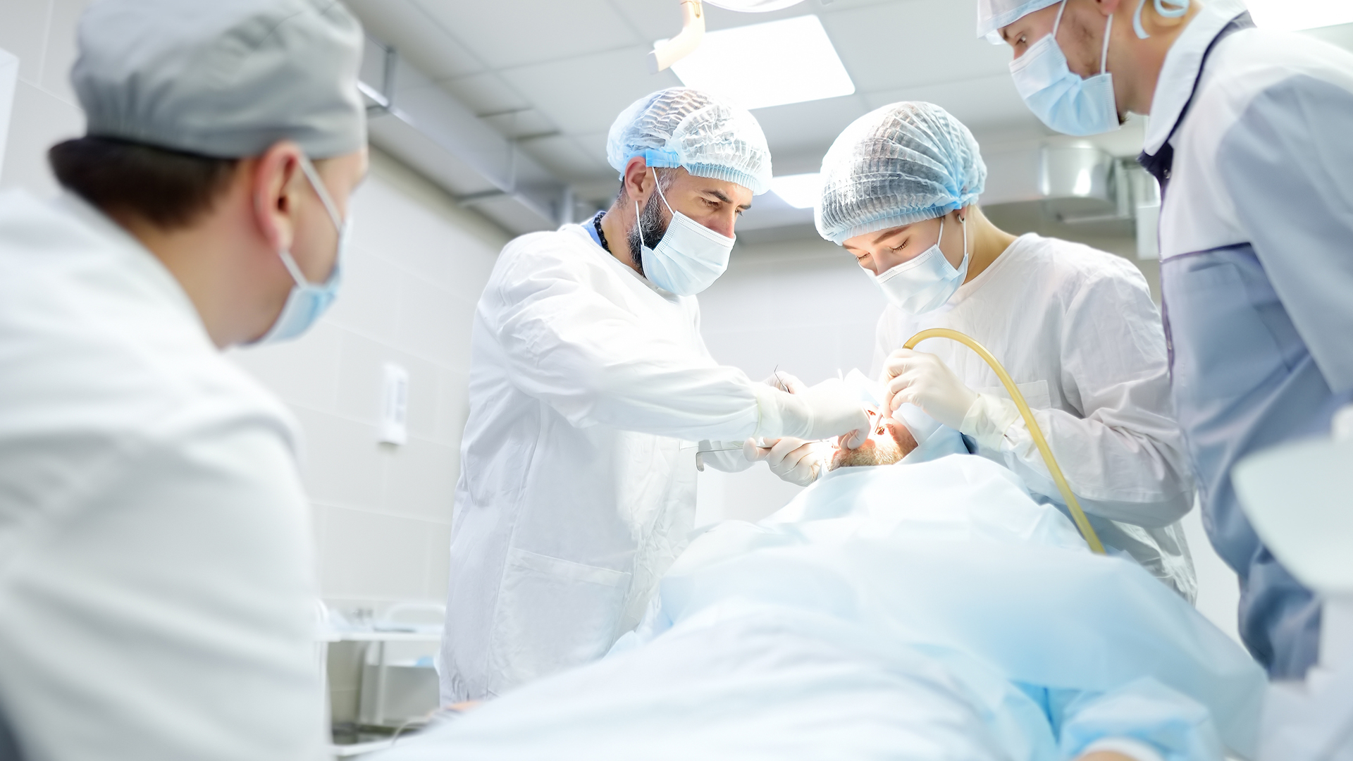 Surgical team in theatre gowns, caps and masks leaning over a patient lying under a surgical drape