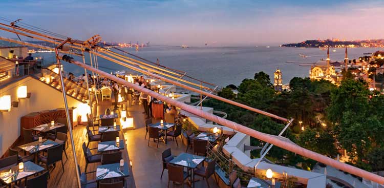 A rooftop terrace at dusk, tables lit by lamps hung from rigging lines, facing the Bosphorus and the Ortaköy mosque
