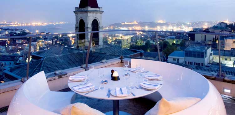 A rooftop restaurant terrace at dusk, a round table laid in front of a stone turret, the roofs of Beyoglu and the old city skyline beyond