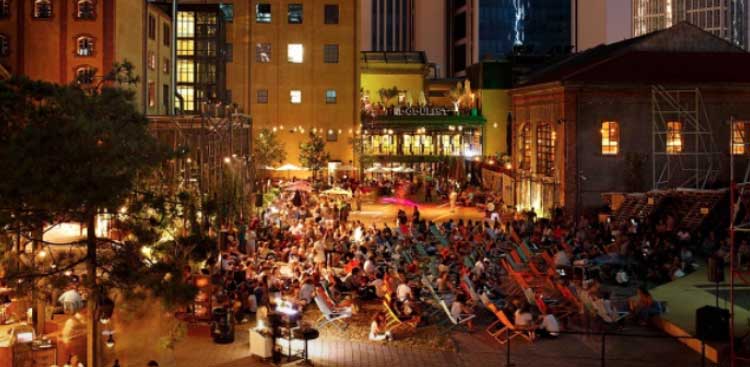The outdoor courtyard of a former brick brewery turned cultural venue, an audience seated in colourful deckchairs in the evening under strings of lights