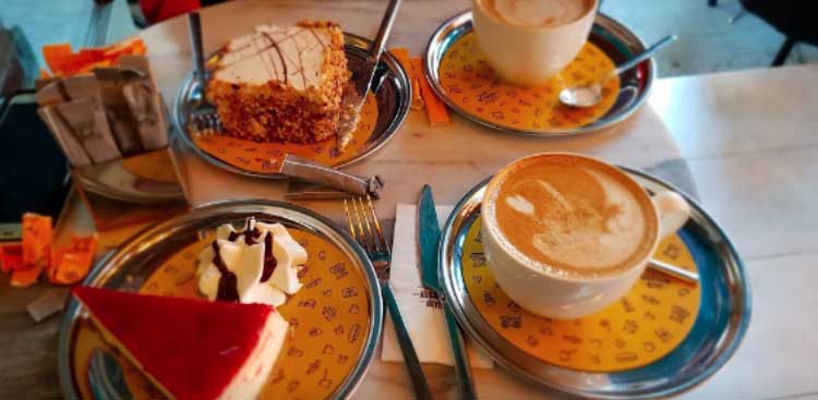 Two cappuccinos and two slices of cake, one with walnuts and one a strawberry cheesecake, served on yellow-patterned metal trays