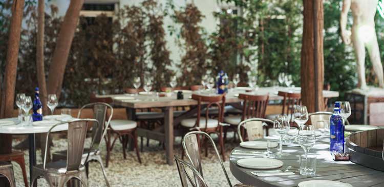 An open-air dining room under the trees, wooden tables and metal chairs standing on gravel, wine glasses and blue bottles on the tables