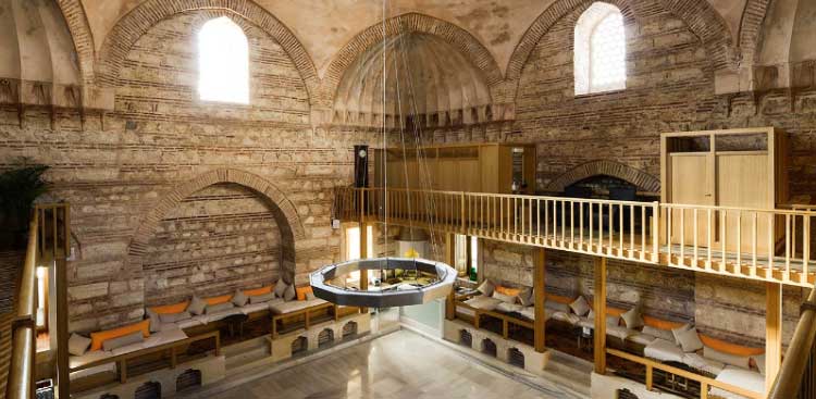 Inside a historic hammam: stone domes and arches, a wooden gallery on the upper floor, cushioned benches around a circular chandelier
