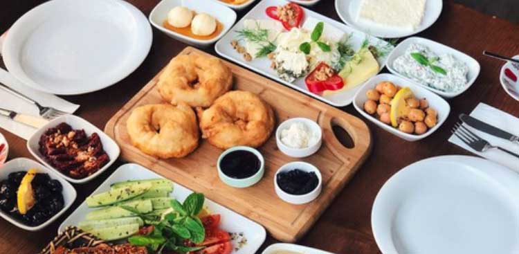 A Turkish breakfast spread: rings of fried dough on a wooden board, cheeses, olives, jams, cucumber and tomato in small dishes