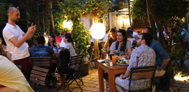 A garden terrace lit by round lamps after dark, guests at tables under the trees in a paved courtyard, one of them taking a photograph