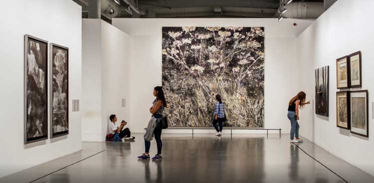 A gallery room in a modern art museum, visitors standing in front of large canvases, among them a dark painting scattered with white flowers