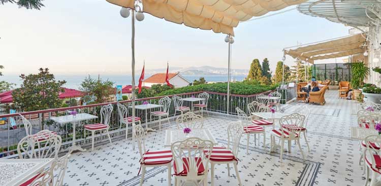The terrace of a Belle Epoque hotel, white wrought-iron furniture and red and white striped cushions on a tiled floor, looking out to sea