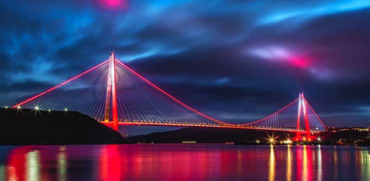 The Yavuz Sultan Selim bridge lit in red above the Bosphorus at night, its reflection stretching across the water