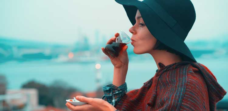 A woman in a wide-brimmed black hat drinking Turkish tea from a tulip glass, the Istanbul skyline and the Bosphorus behind her
