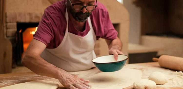 A baker in an apron stretching dough on a floured worktop in front of a lit wood-fired oven