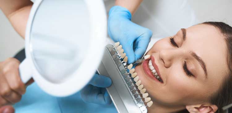 Patiente allongée au fauteuil tenant un miroir pendant que la praticienne compare un teintier à ses dents