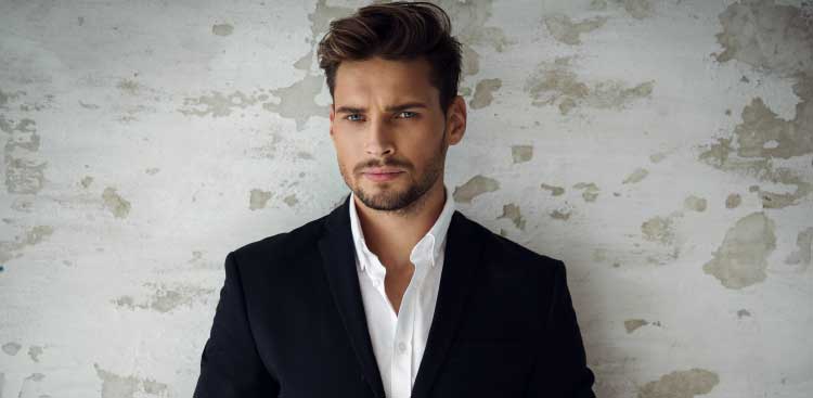 Man wearing a dark jacket and white shirt against a pale wall