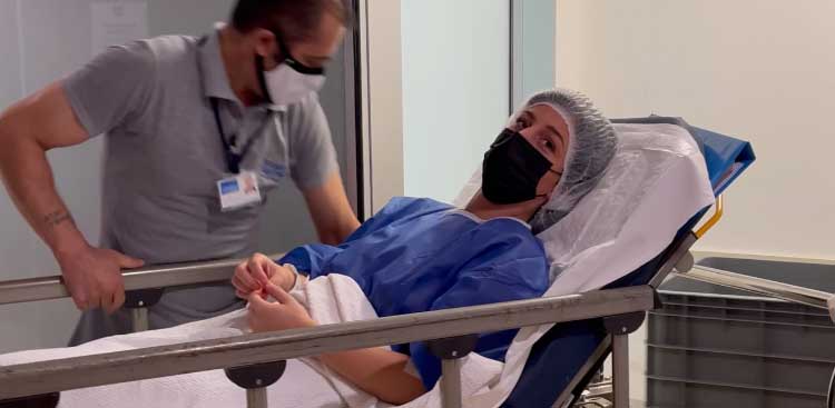 Océanie lying on a trolley, in a surgical cap and theatre gown, being wheeled by a member of staff along a corridor in the clinic