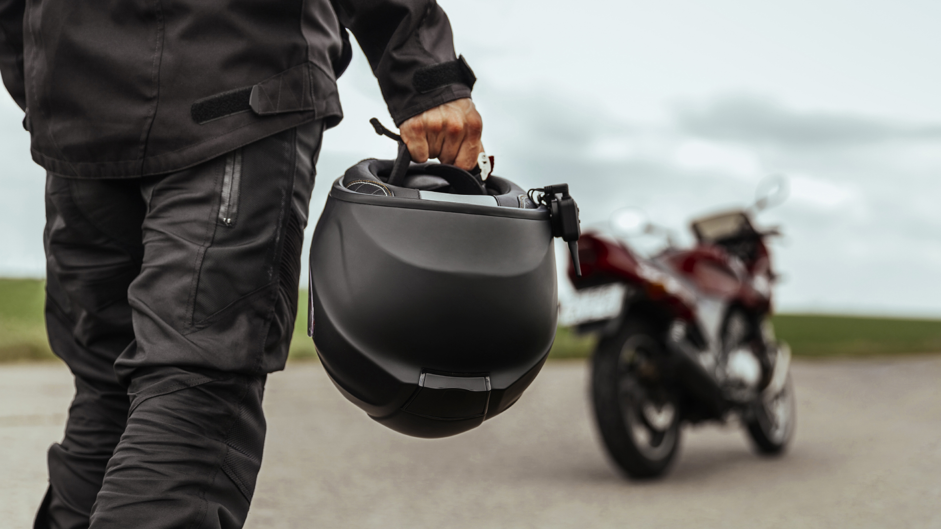 Personne en tenue de moto noire tenant un casque intégral noir, avec une moto rouge floue à l’arrière-plan.