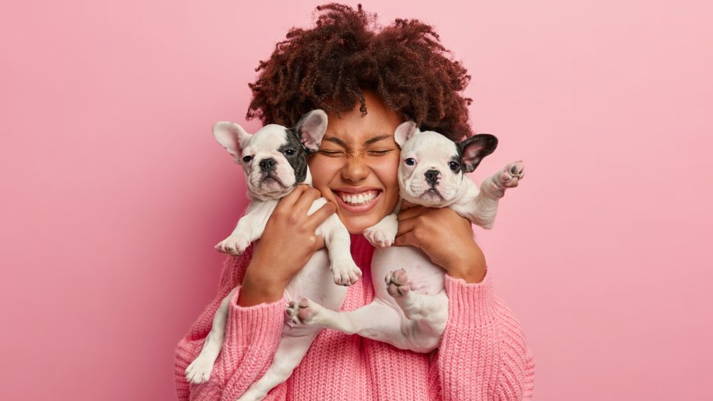 Une femme aux cheveux bouclés sourit les yeux fermés en serrant contre elle deux chiots bouledogues français, sur fond rose