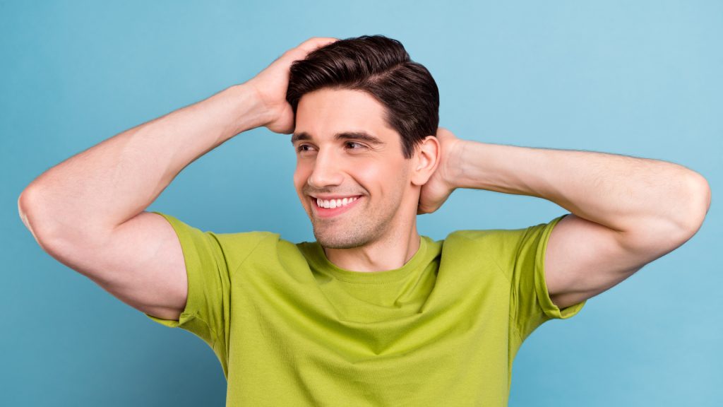 Un homme en t-shirt vert passe les deux mains dans ses cheveux en souriant, sur fond bleu