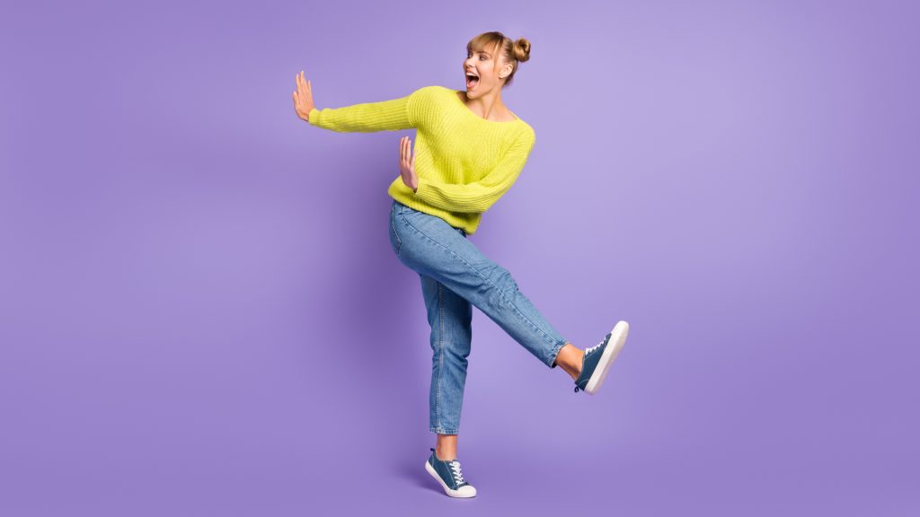Une femme en pull jaune et jean s'élance une jambe levée et les bras écartés, sur fond violet