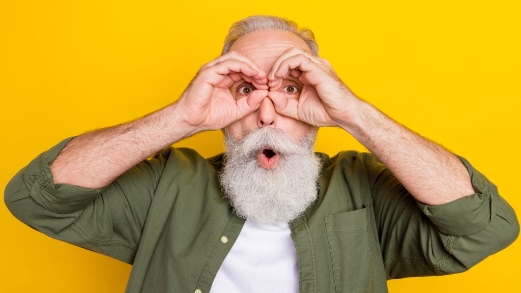 Un homme âgé à barbe blanche forme des jumelles avec ses mains devant ses yeux, sur fond jaune
