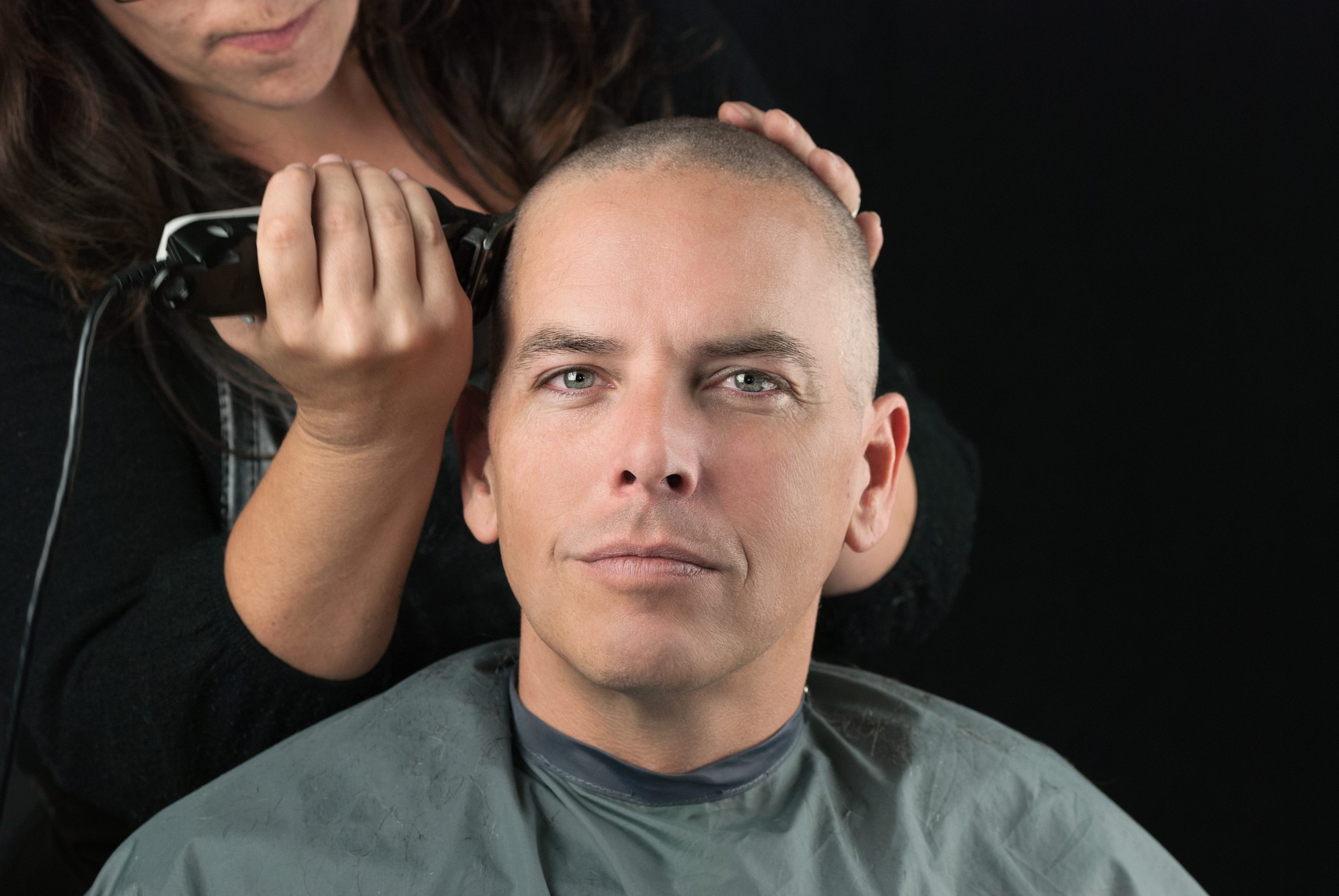 Une personne passe une tondeuse sur le côté du crâne d’un homme aux cheveux très courts, en maintenant sa tête de l’autre main.