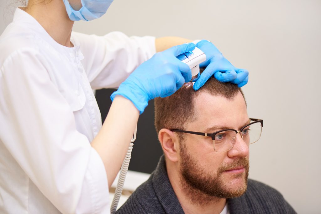 Un professionnel portant des gants bleus examine le cuir chevelu d’un homme avec un appareil optique portatif.