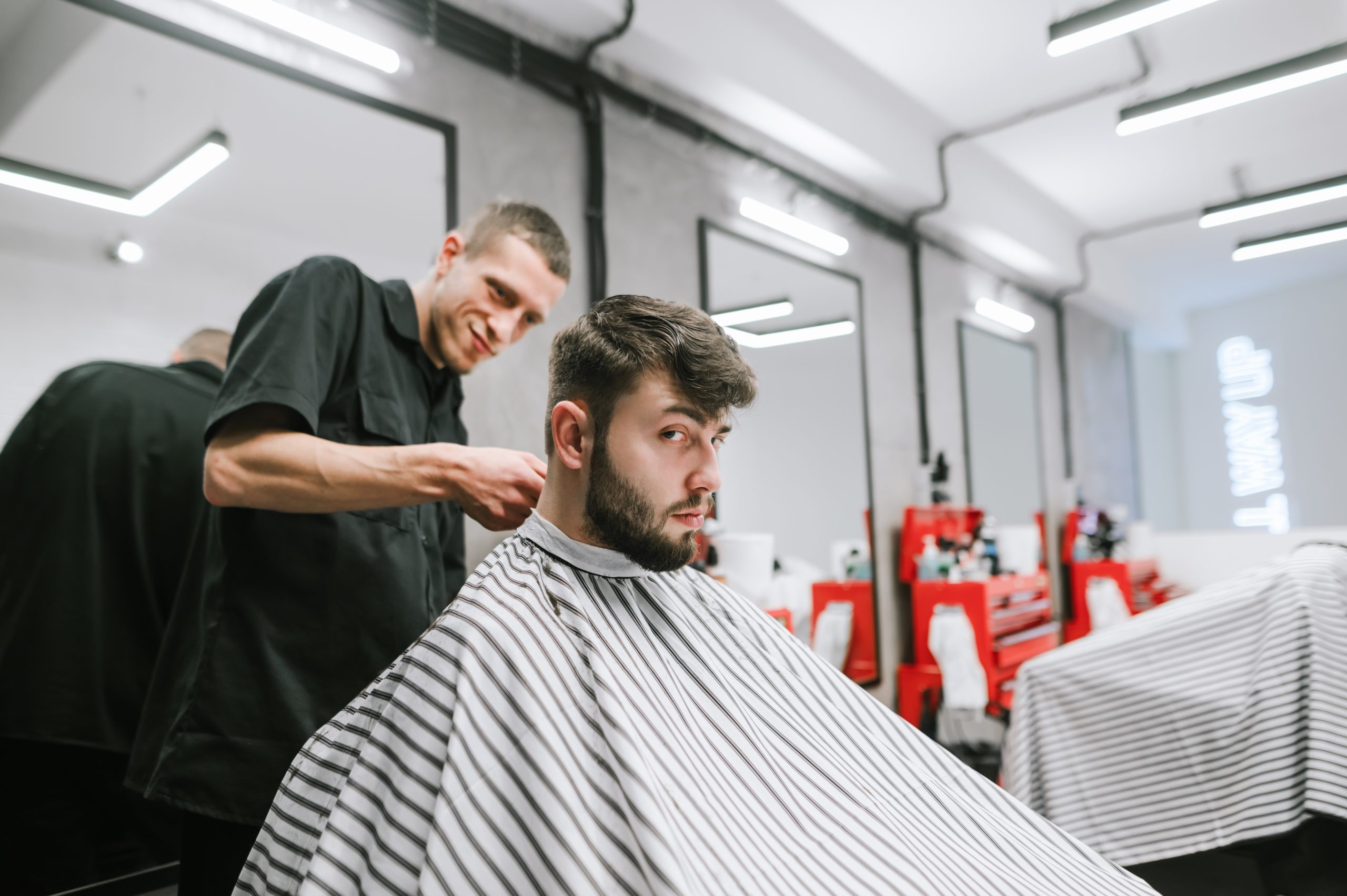 Un coiffeur travaille à l’arrière de la tête d’un homme assis sous une cape rayée dans un salon.