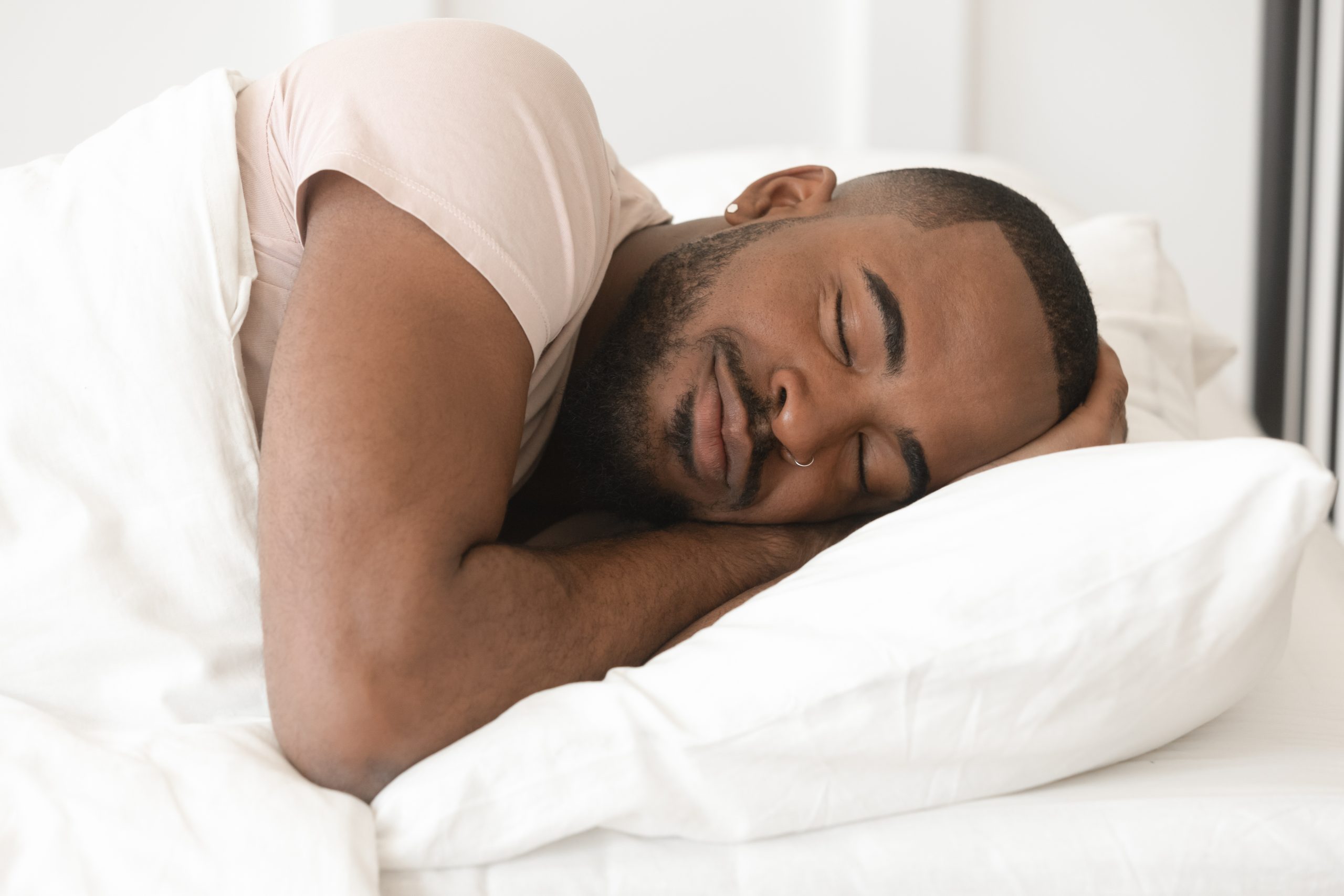 Man sleeping on his side, his head resting on a white pillow and one arm bent near his face.