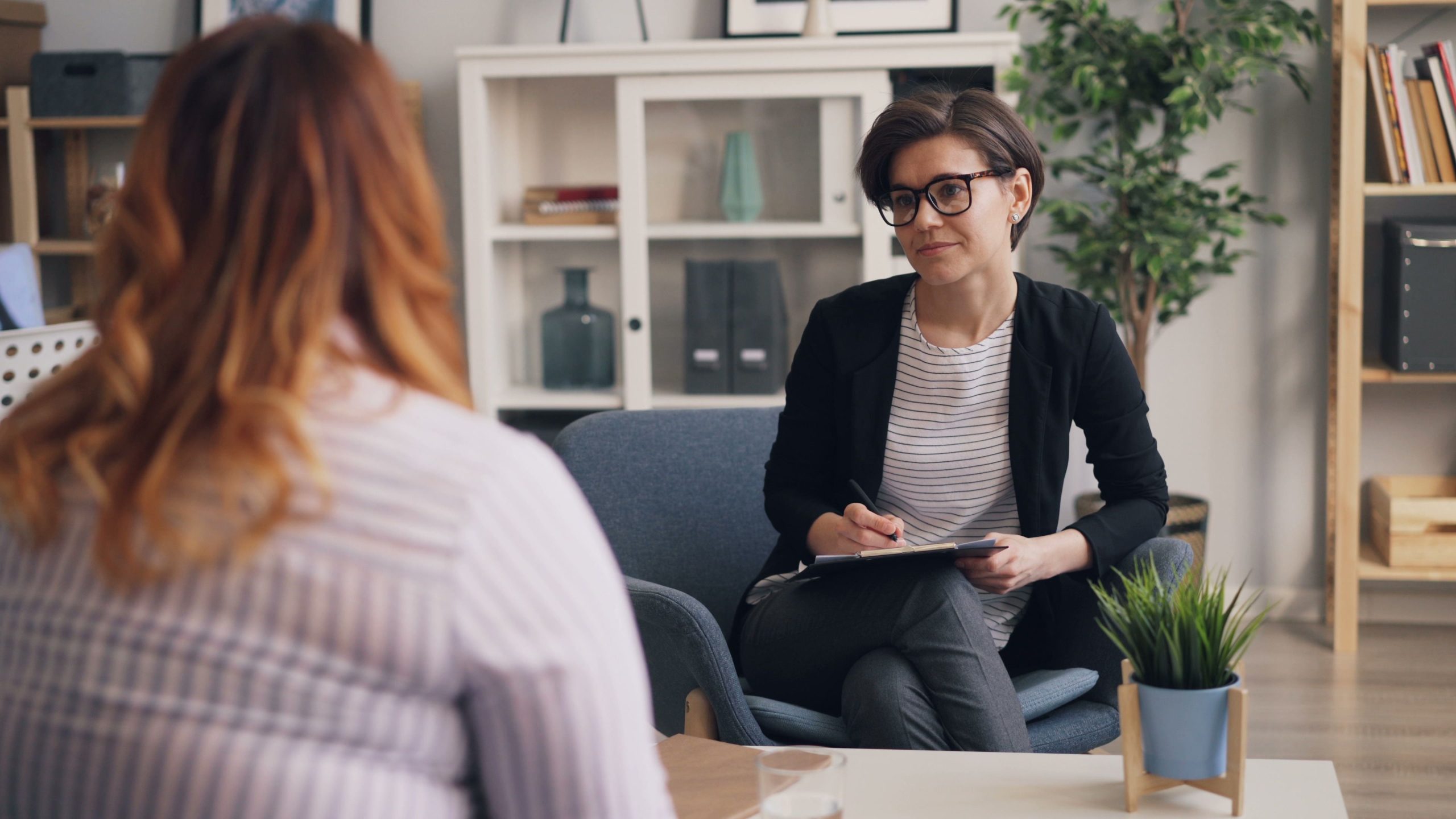 A woman wearing glasses holds a pen and clipboard opposite another woman seen from behind.