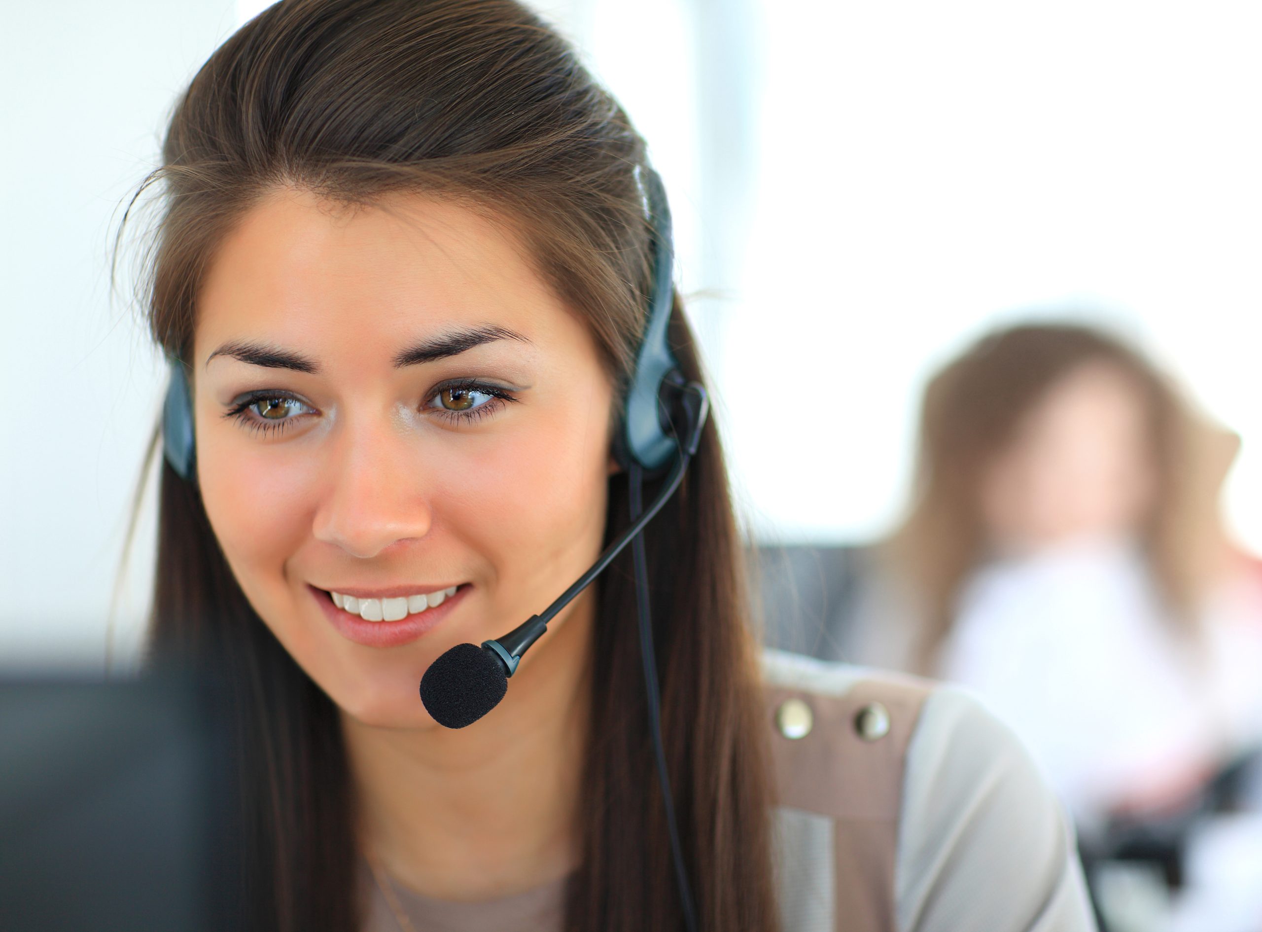 Smiling woman wearing a headset with a microphone in an office, with a blurred person in the background.