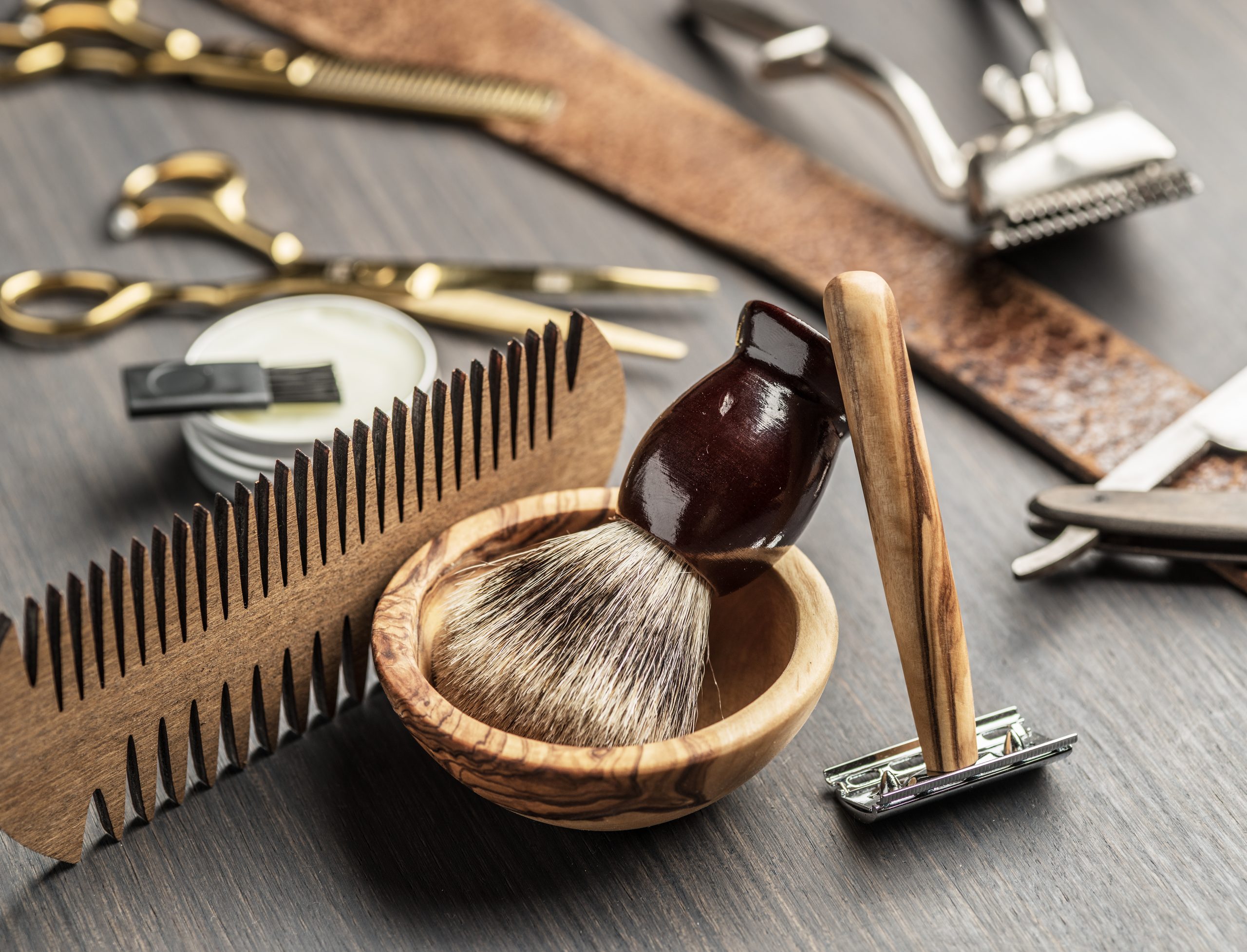 Peigne, ciseaux, rasoirs et blaireau dans un bol en bois, disposés sur une table.