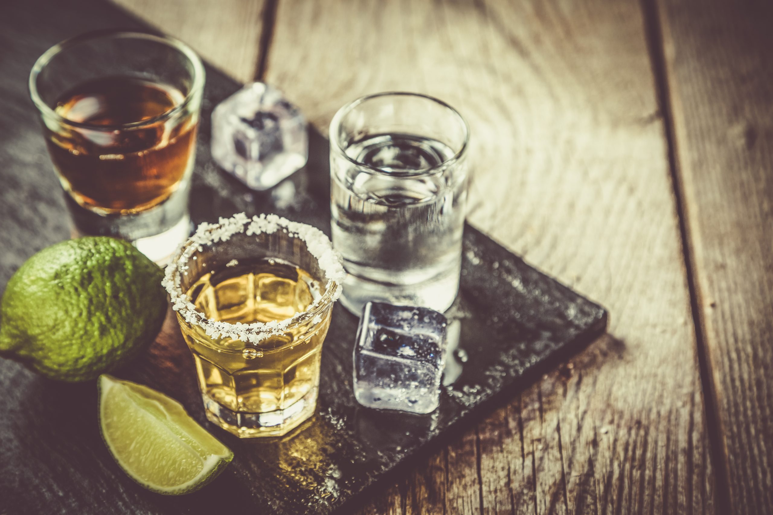 Three small glasses of amber or clear drinks, with lime, salt and ice cubes on a wooden table.