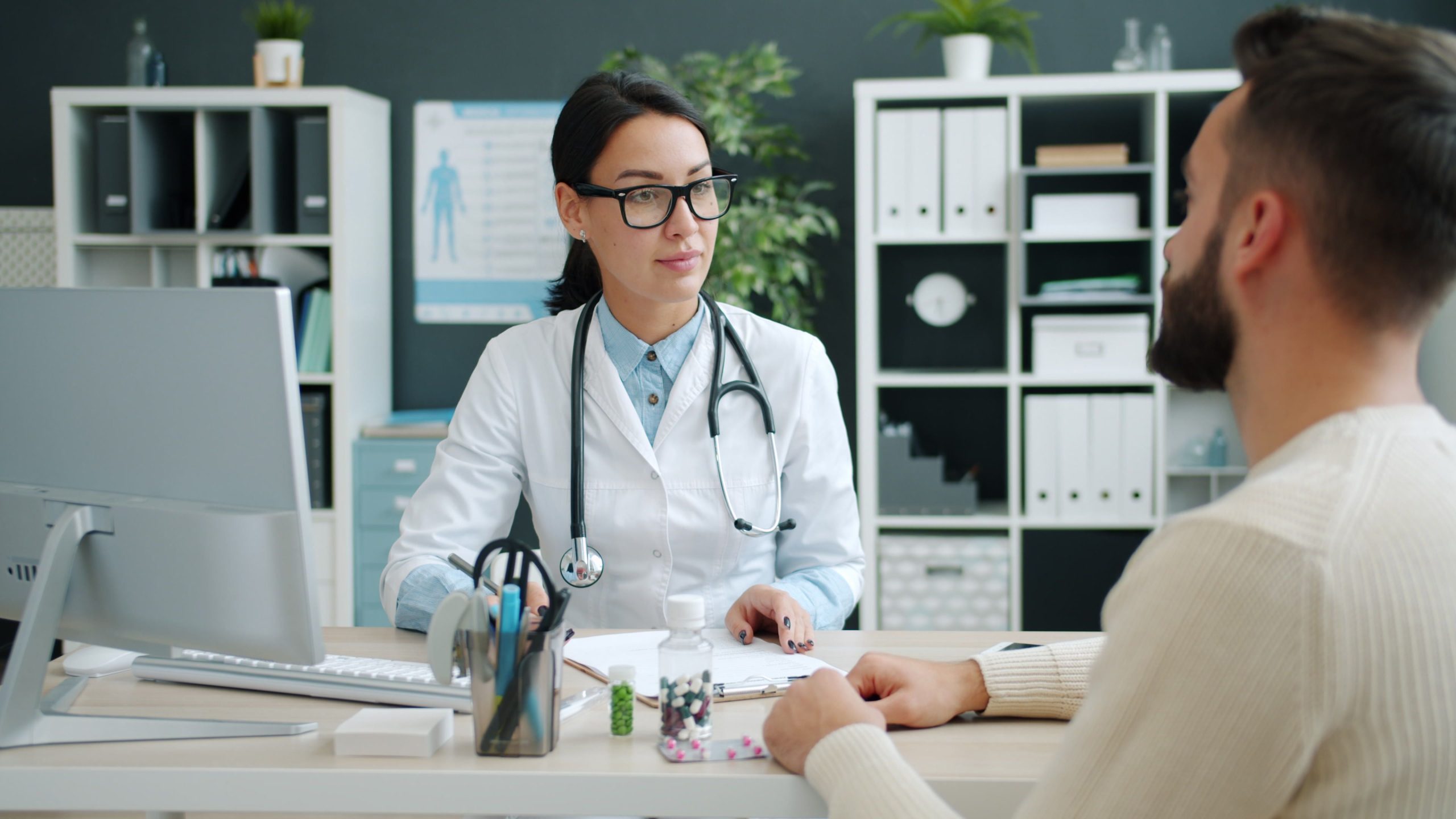 A woman in a white coat with a stethoscope talks with a man, with bottles of tablets on the desk.
