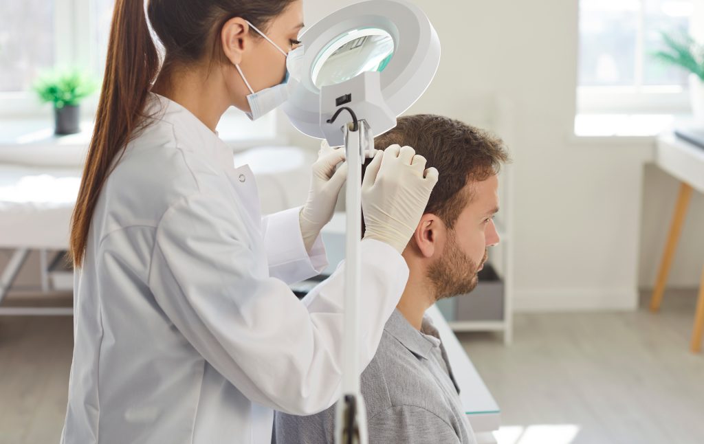 A masked professional wearing gloves examines a seated man’s scalp using a magnifying lamp.