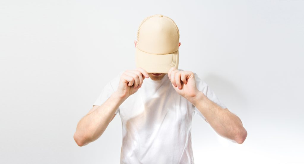 Homme en tee-shirt blanc, tête baissée, tenant des deux mains la visière de sa casquette beige.