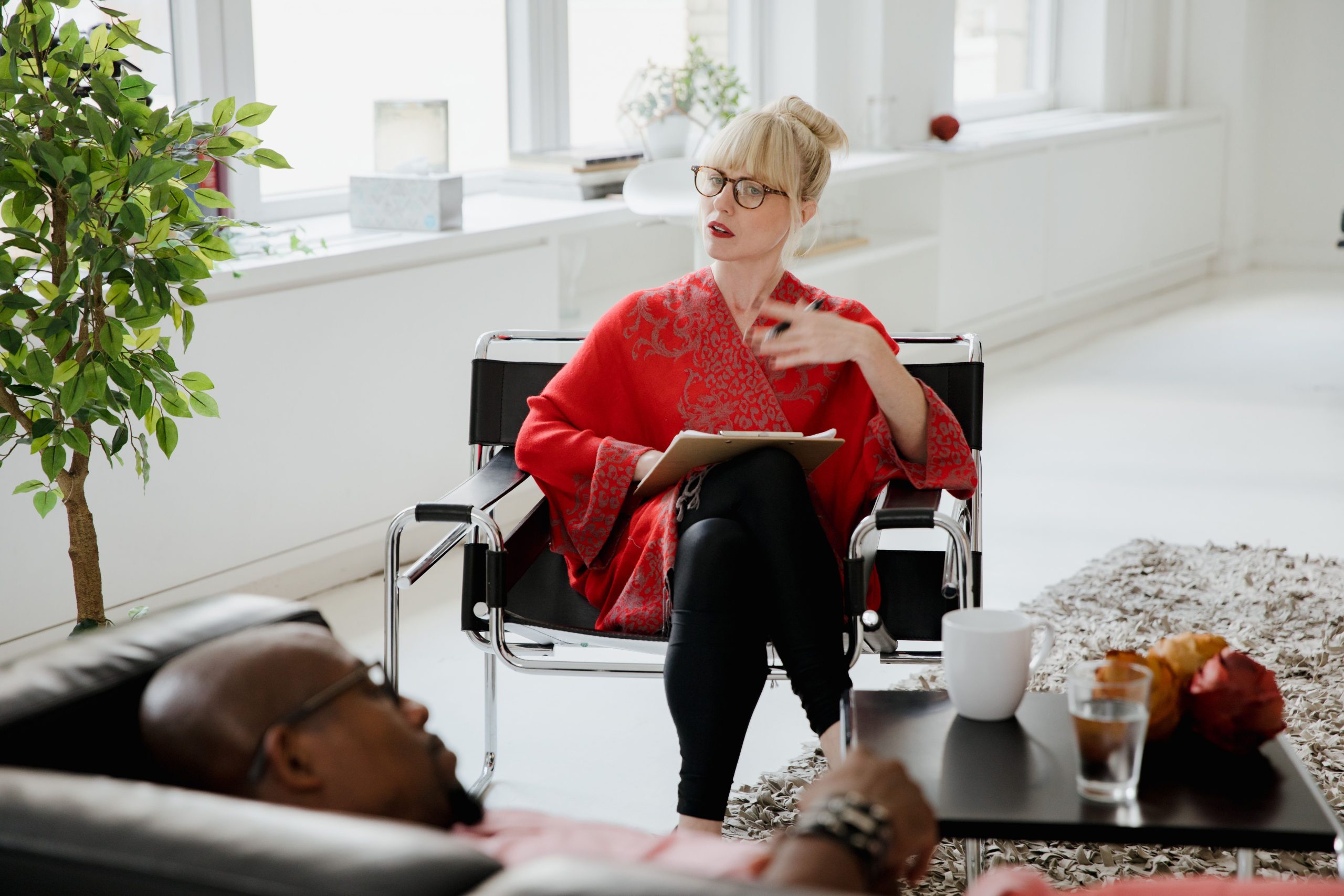 A woman with a clipboard on her lap talks with a man reclining in an armchair in a bright room.