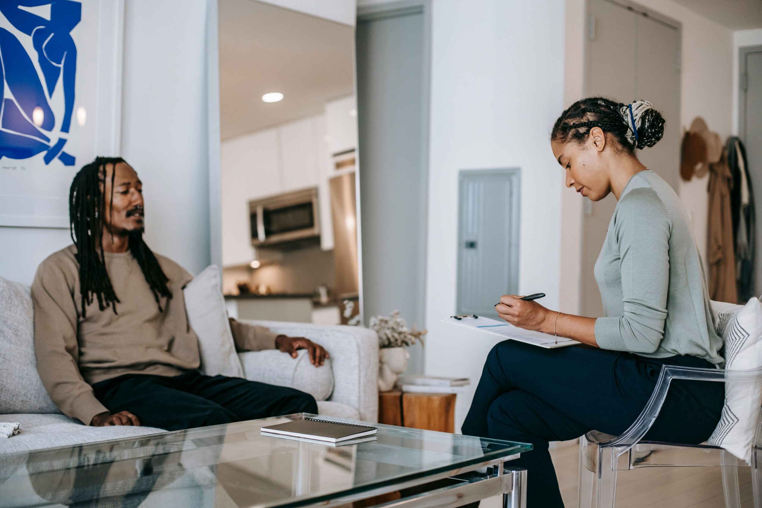 A woman takes notes on a clipboard opposite a man seated on a sofa.
