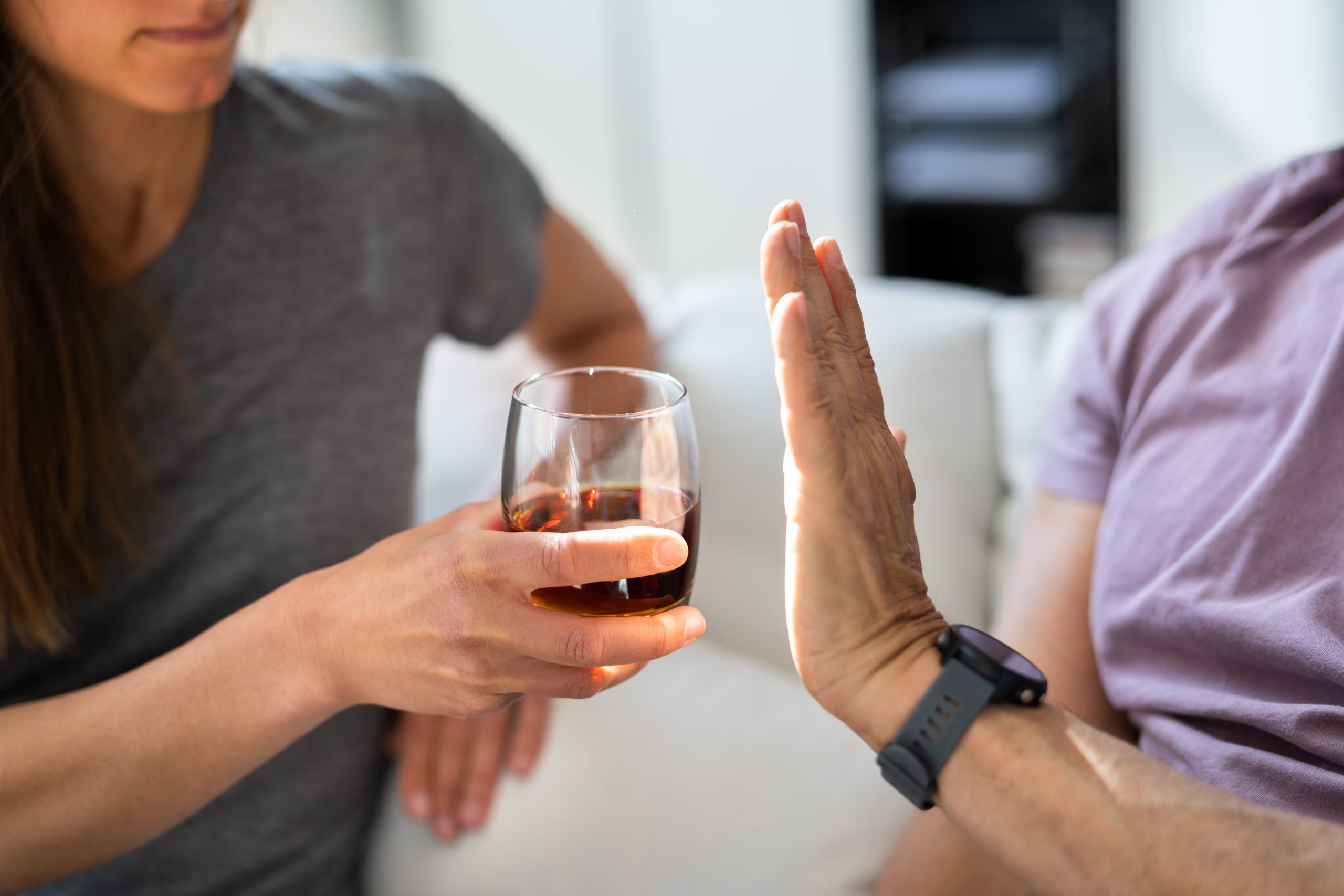 A person raises a hand to refuse a glass of amber liquid offered by another person.
