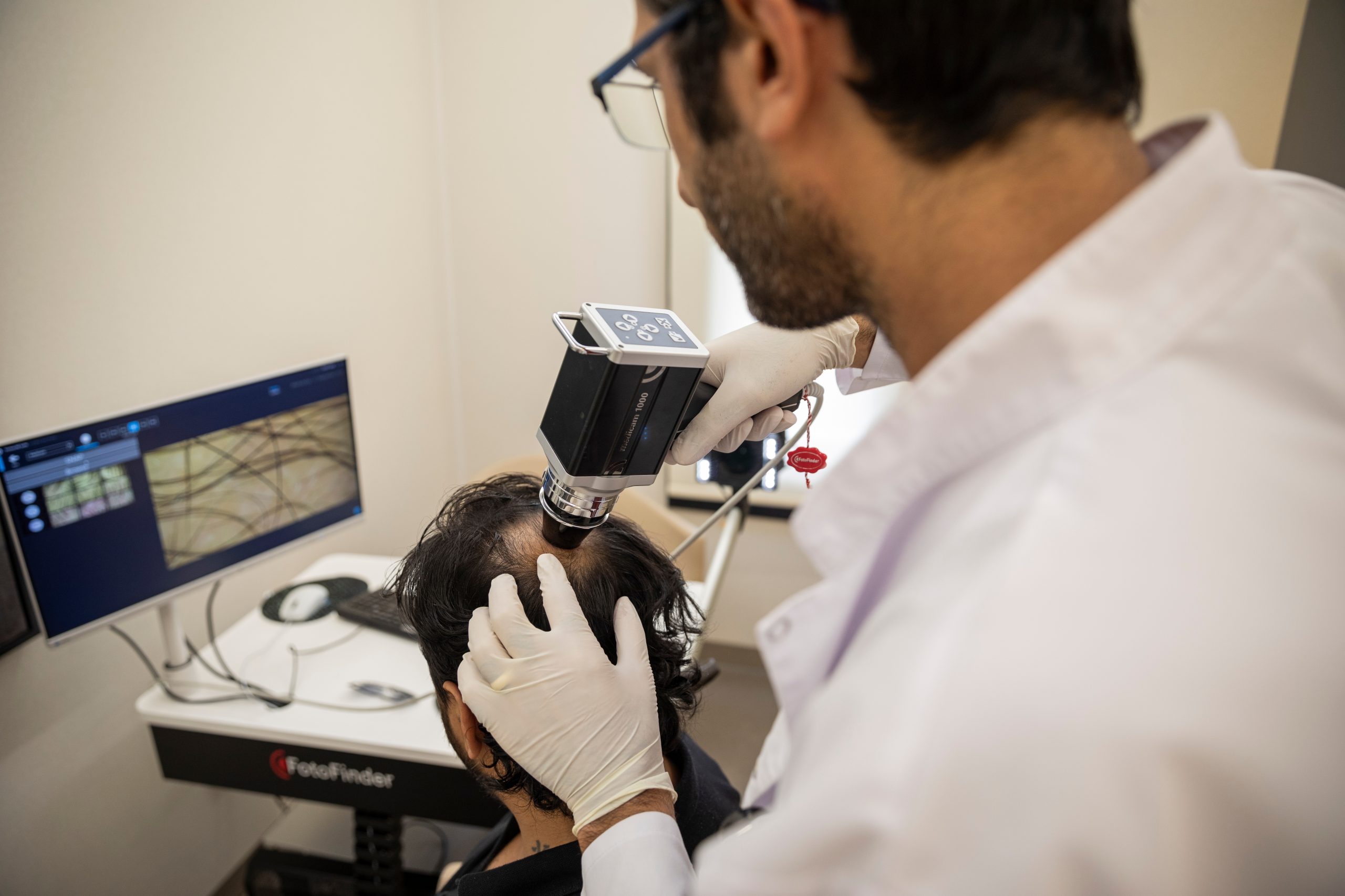 Un praticien ganté écarte les cheveux et examine le cuir chevelu avec un appareil optique, tandis qu’un moniteur affiche les cheveux grossis.