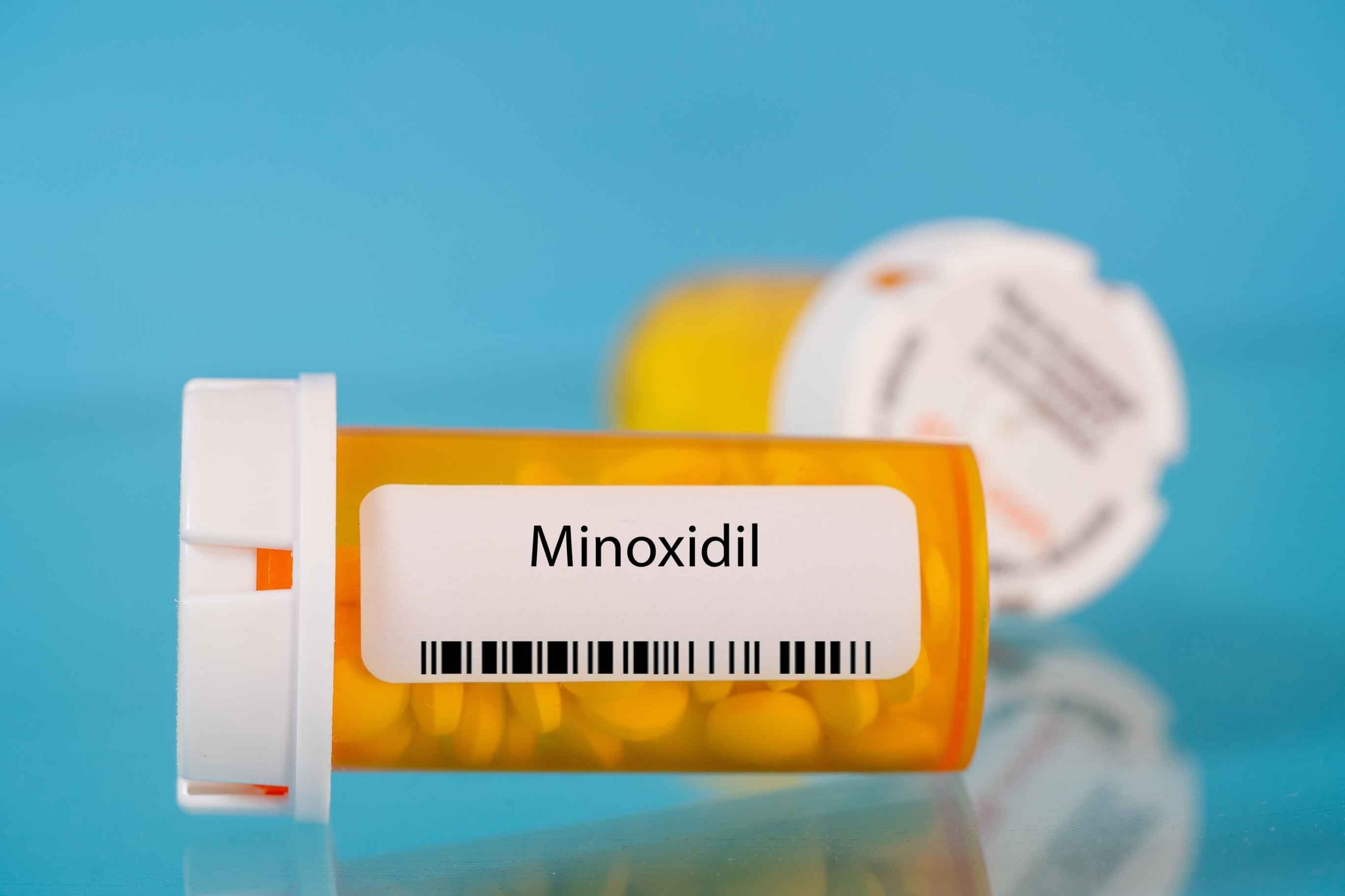 Orange bottle labeled “Minoxidil,” containing yellow tablets, lying on a blue background.