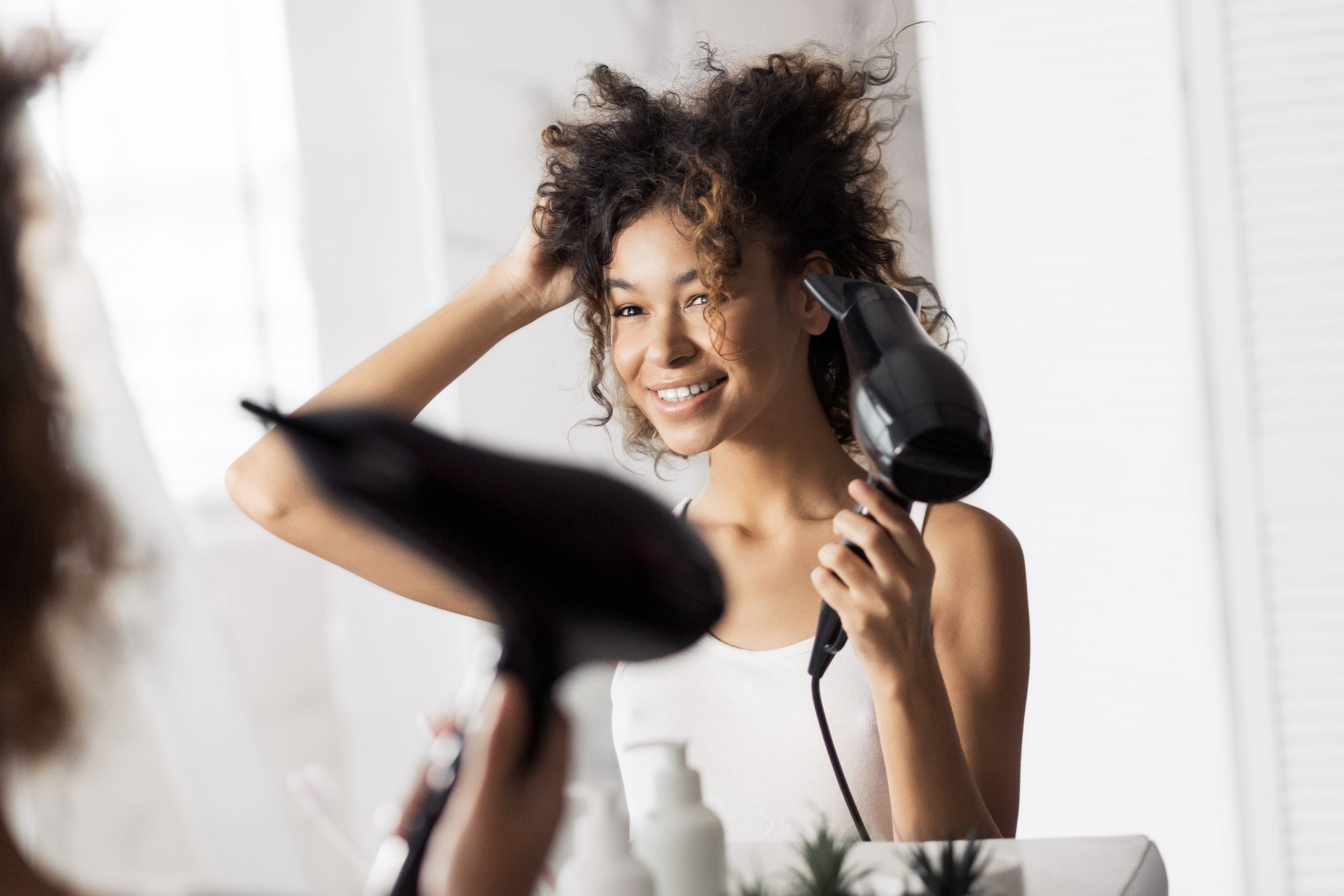 Une femme sèche ses cheveux bouclés avec un sèche-cheveux devant un miroir.